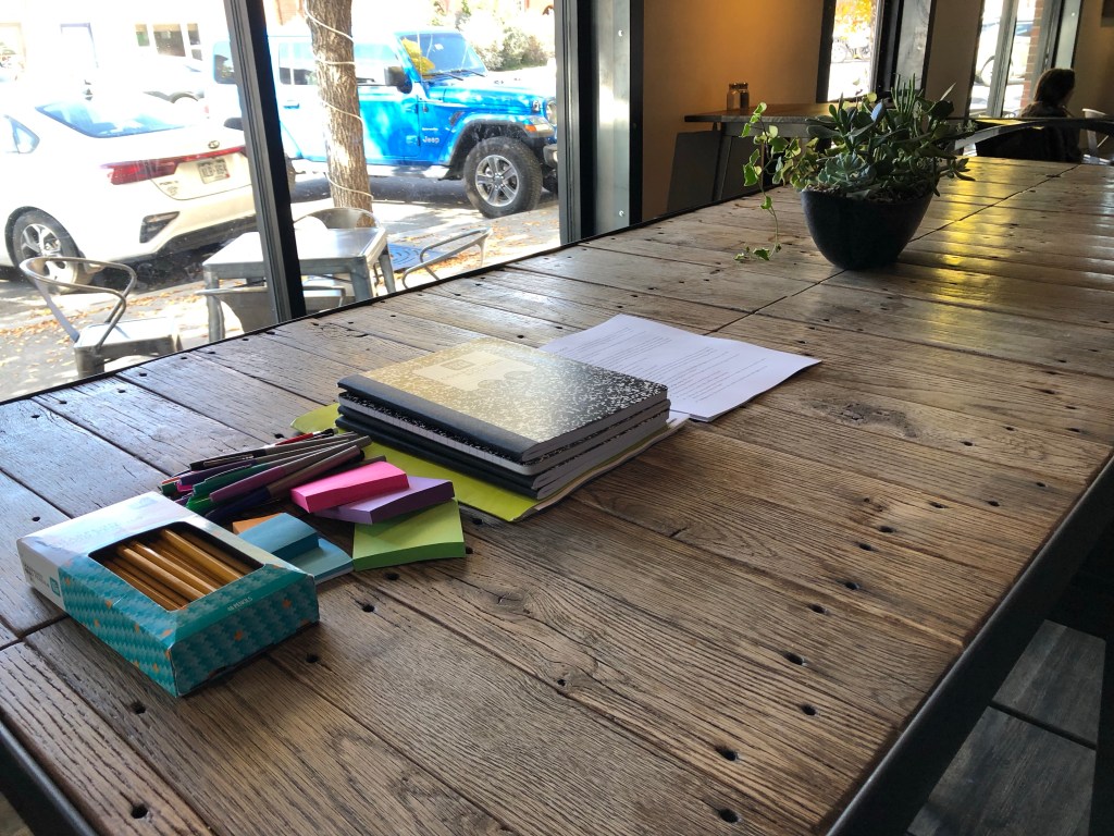 a long table with a stack of notebooks, sticky notes, and pencils organized in a row. A plant sits on the right edge of the frame.