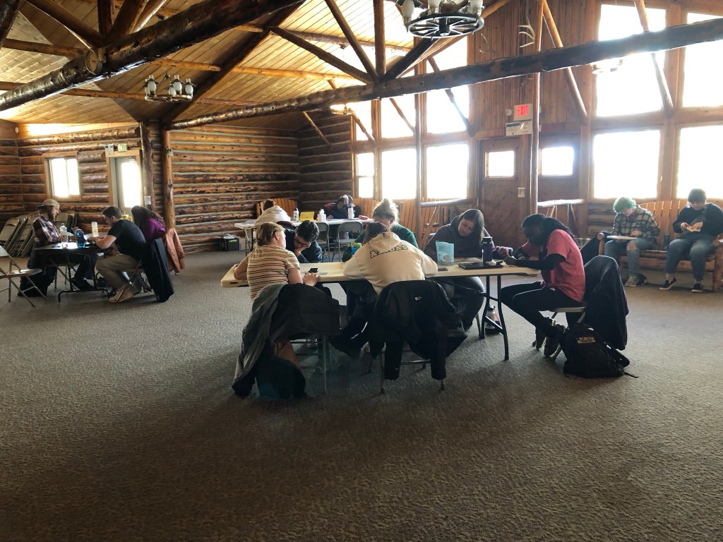 several people sit around different tables in a large room with a high ceiling. The ceiling has wooden beams, and light coming in through the windows.
