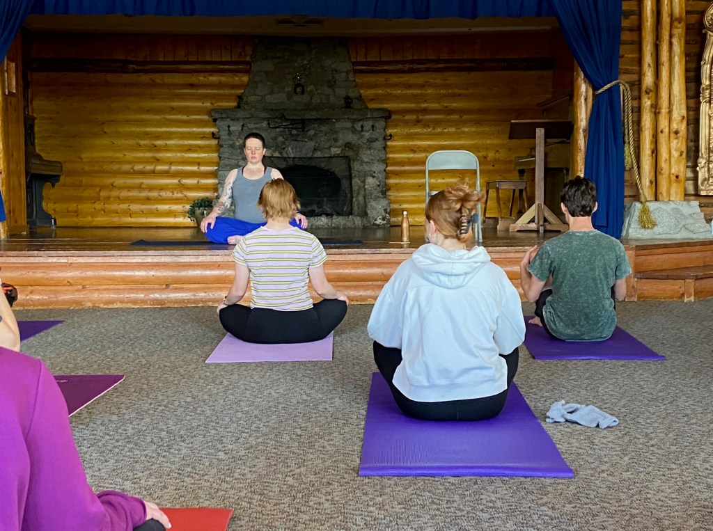 a person with short hair, wearing blue pants and a gray shirt, sits on a stage with eyes closed. 3 people sit on the floor facing her. 