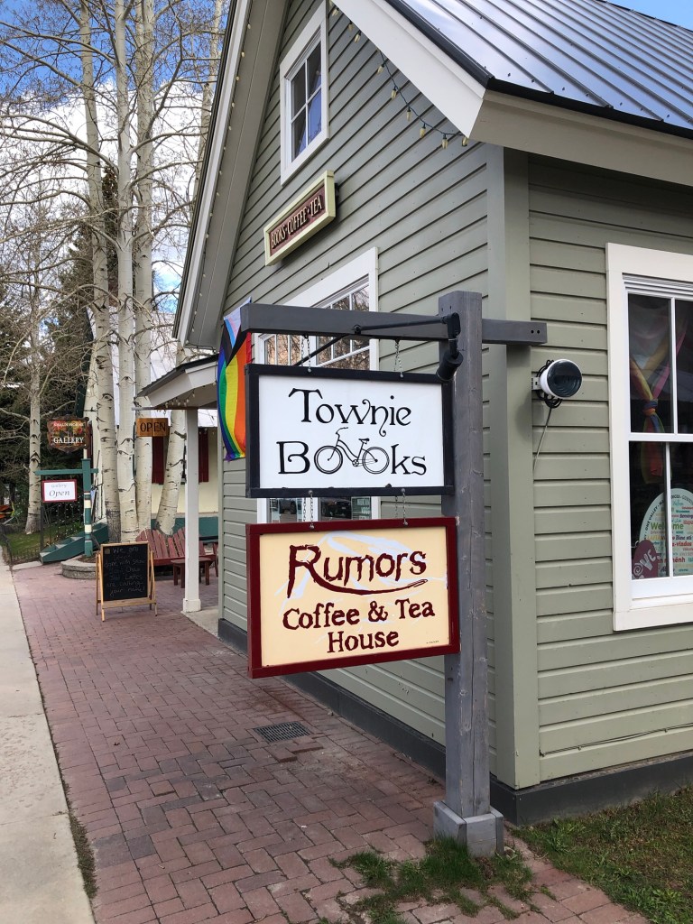 image of a double hanging sign outside a green building. The top sign reads "Townie Books" and the bottom sign reads "Rumors Coffee and Tea House" 